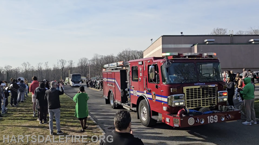 A fire truck on a road with people gathered, some waving, likely celebrating an event or procession.