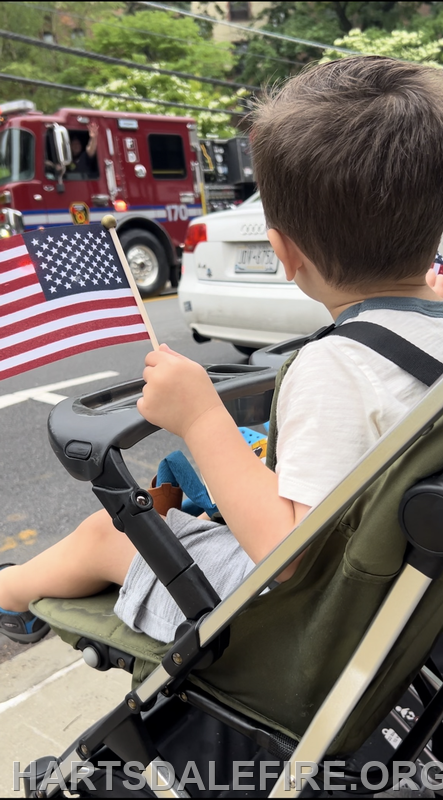 A child holding a small American flag sits in a stroller, watching a vehicle in a parade or procession.