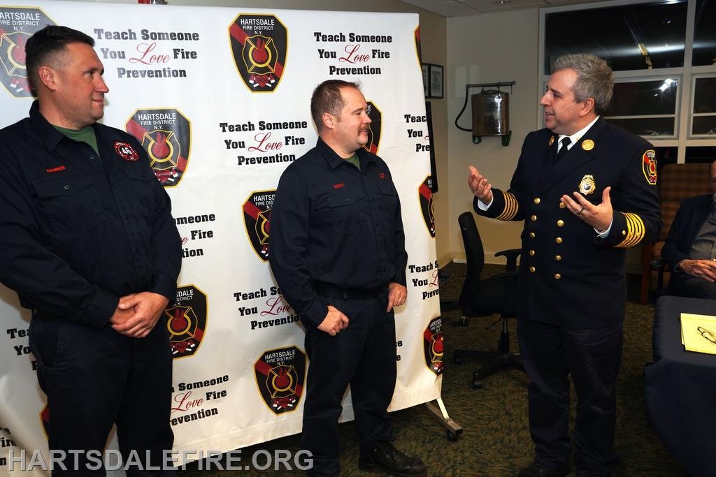 Firefighters and a chief discuss fire prevention, with a backdrop promoting fire safety education in a meeting setting.
