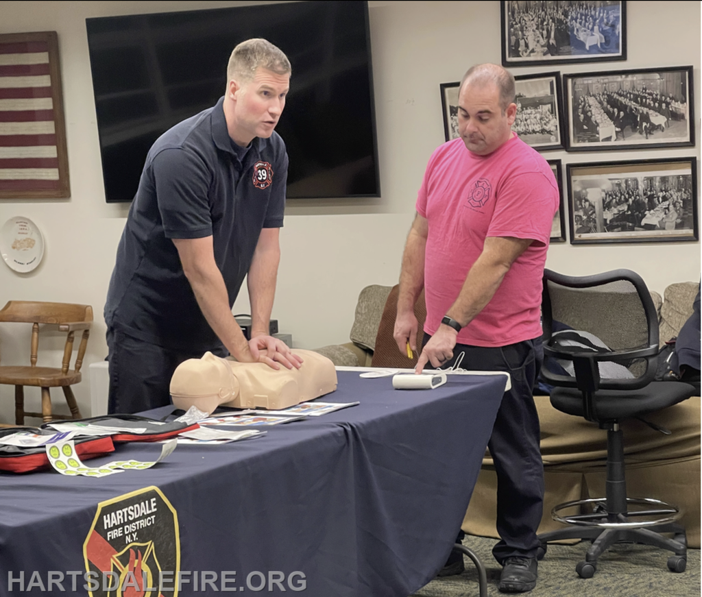 Two instructors demonstrate CPR techniques on a mannequin during a training session. Learning materials are visible on the table.