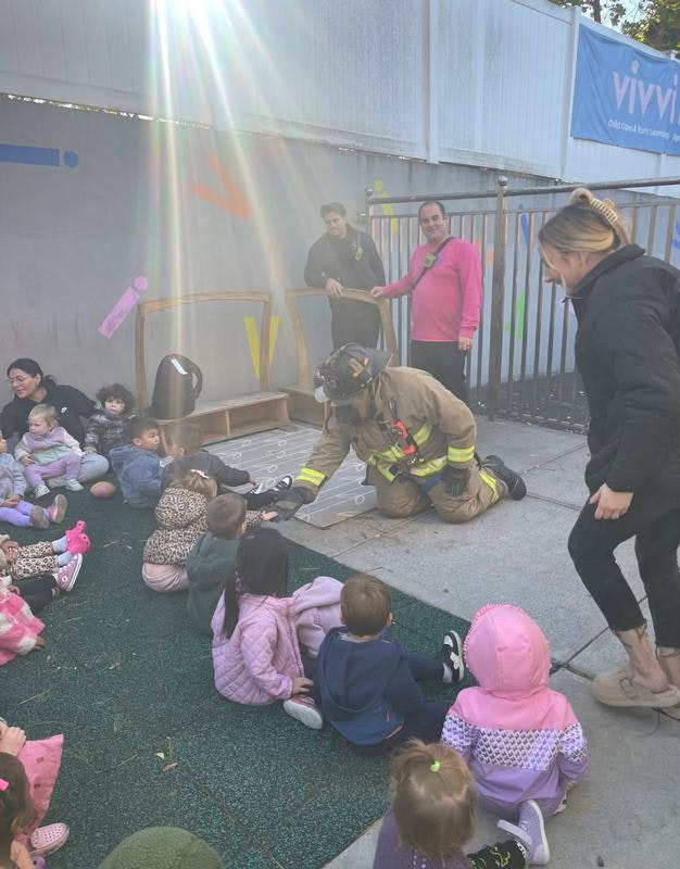 A firefighter interacts with children during a safety demonstration, while adults observe nearby in a playful, colorful setting.