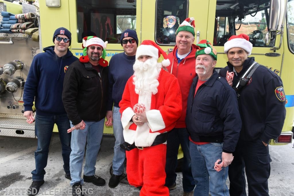 A group of people in holiday attire, including Santa and elves, standing by a fire truck with candy canes.