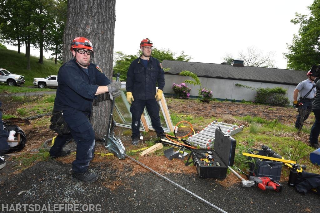 Firefighters in protective gear are working outside with tools and equipment, near a ladder and toolbox, next to a tree.
