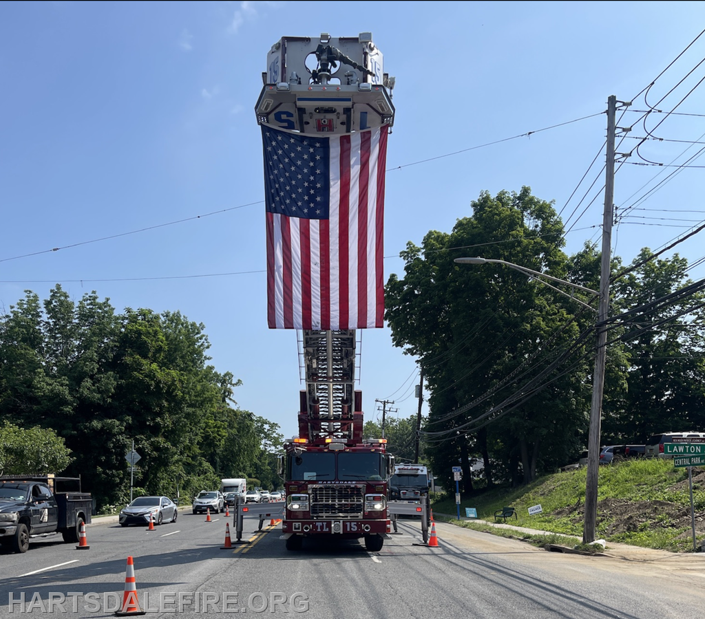 A fire truck with an extended ladder flying an American flag on a busy road, surrounded by traffic and construction cones.