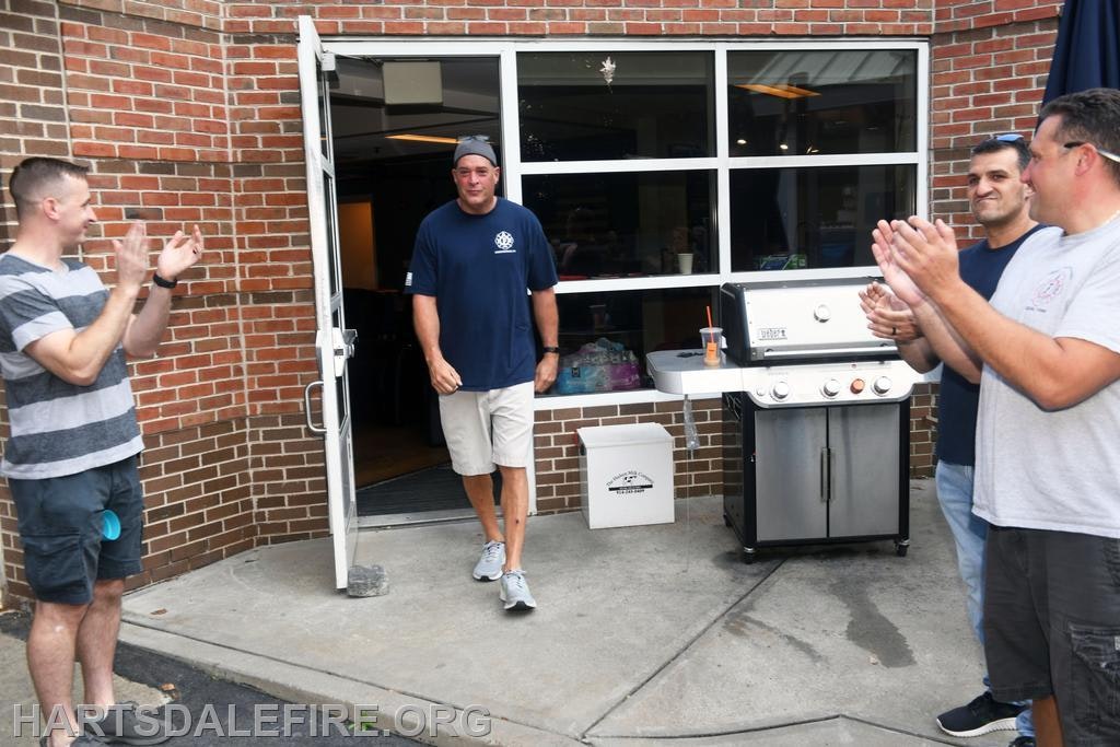 A group of men applaud as one walks out from a building, near a grill, suggesting a celebration or gathering event.
