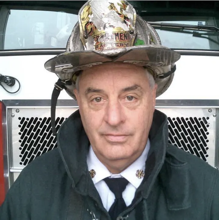 A firefighter in uniform, wearing a helmet, stands in front of a fire truck, showing a serious expression.