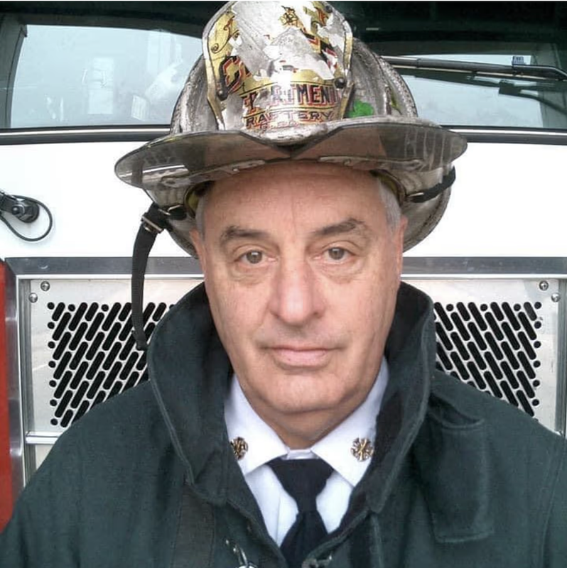 A firefighter in uniform, wearing a helmet, stands in front of a fire truck, showing a serious expression.