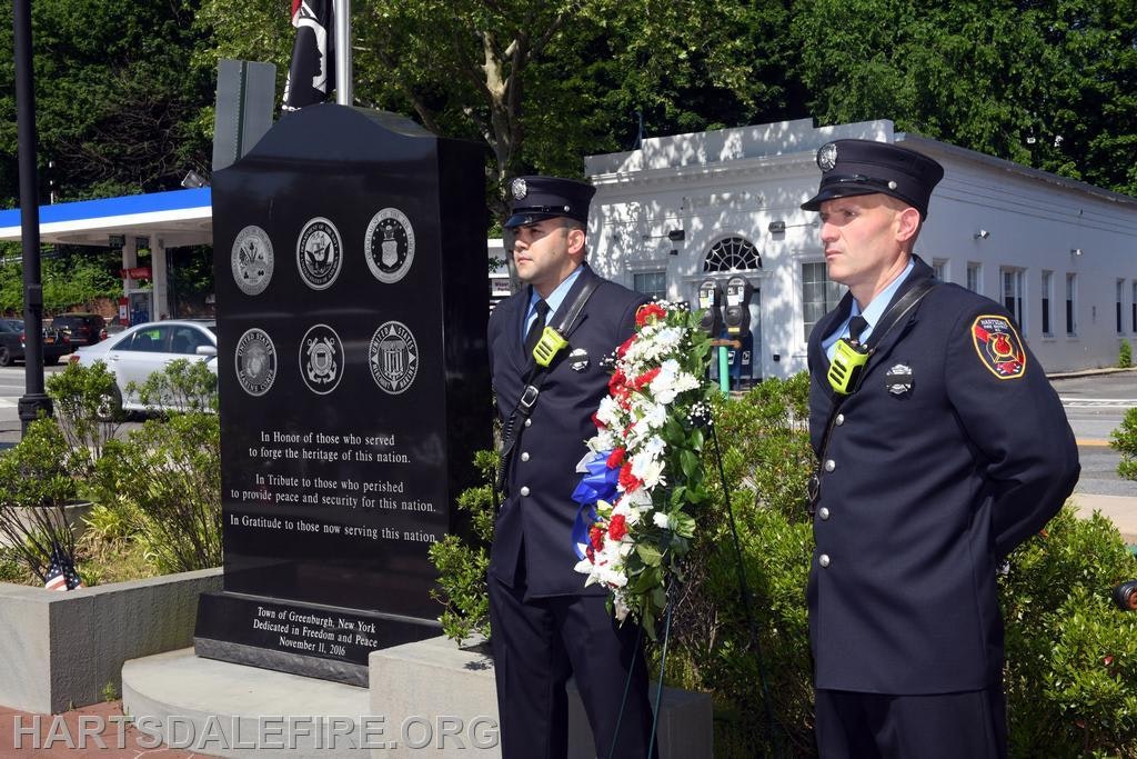 Two uniformed individuals stand by a memorial monument with military insignia and a wreath.