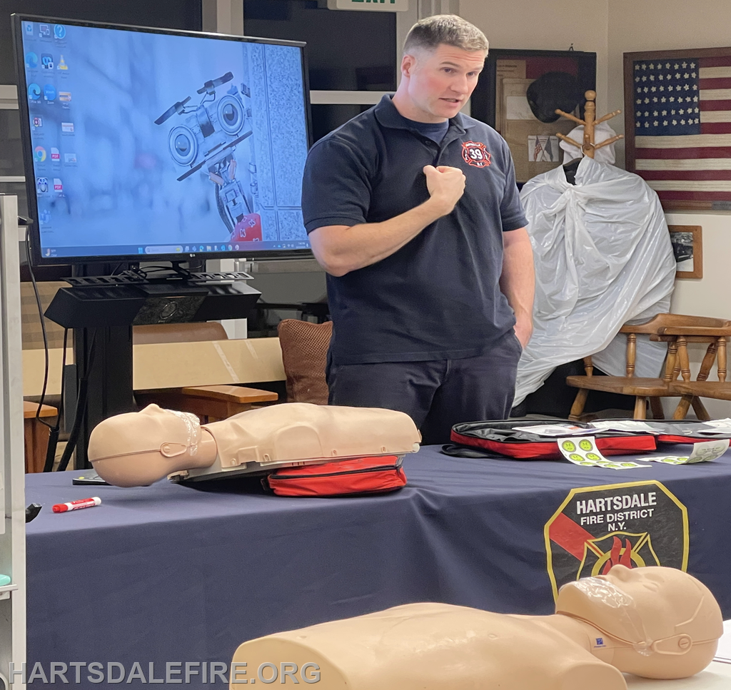 A firefighter is teaching a class with mannequins for CPR practice, alongside a presentation screen in the background.