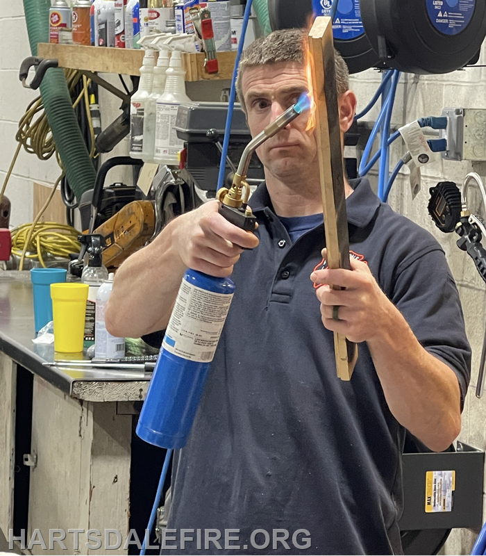 A person is holding a blowtorch, igniting a piece of wood, in a workshop setting filled with various tools.