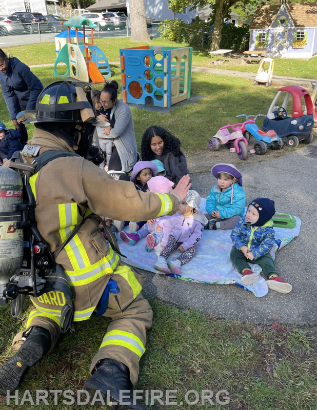 A firefighter interacts with a group of young children in a playground setting, while adults observe nearby.