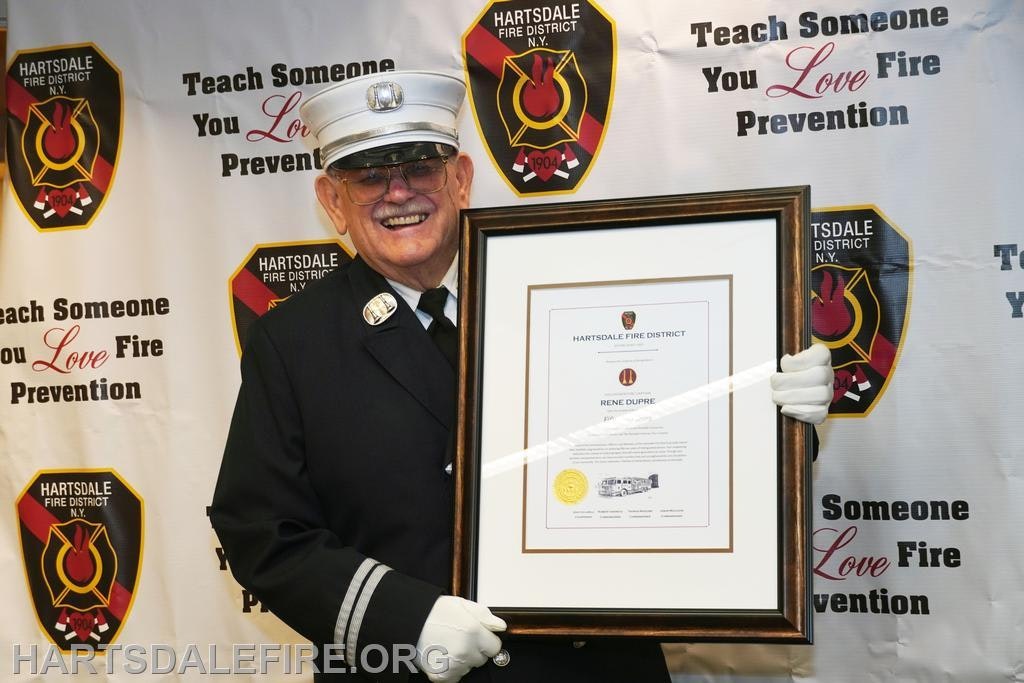 A smiling man in a fire department uniform holds a framed certificate against a backdrop promoting fire prevention education.
