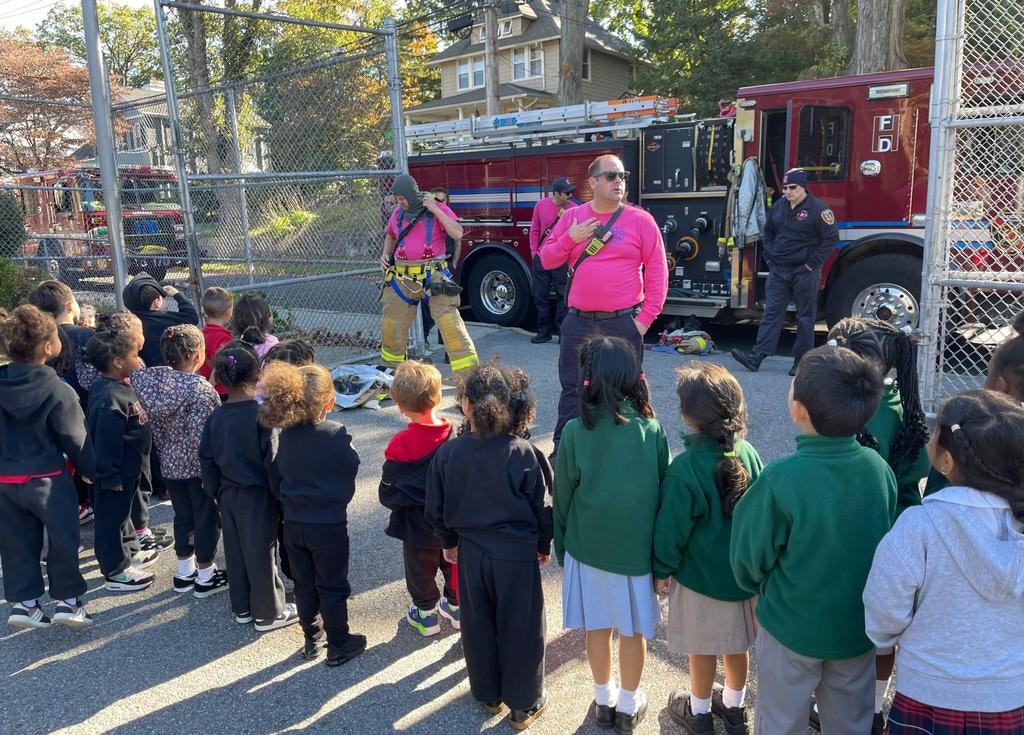 A group of young children watches as firefighters, in pink uniforms, demonstrate safety procedures near a fire truck.