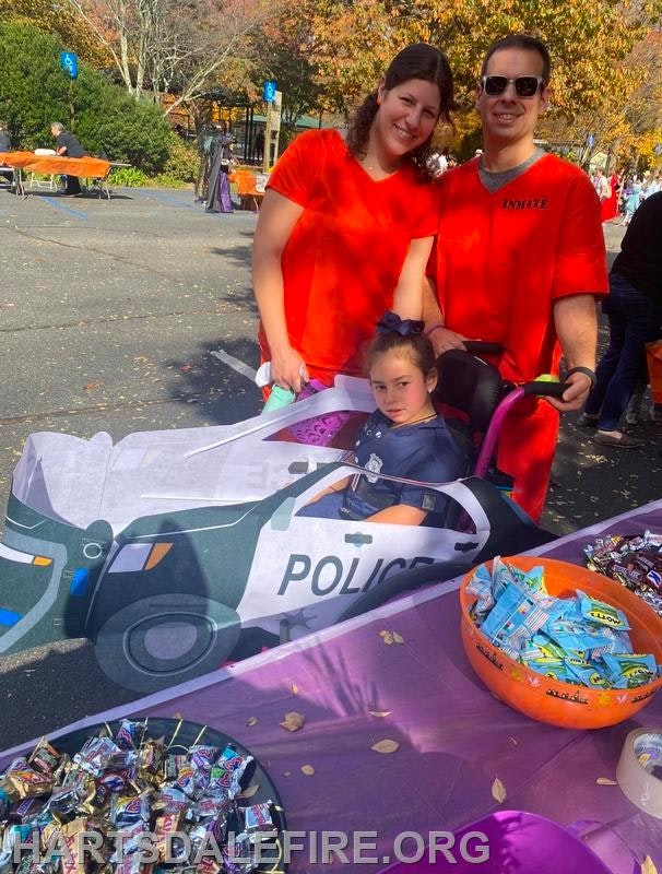 A family is at a festive event, with a girl in a decorated stroller resembling a police car, surrounded by candy on a table.