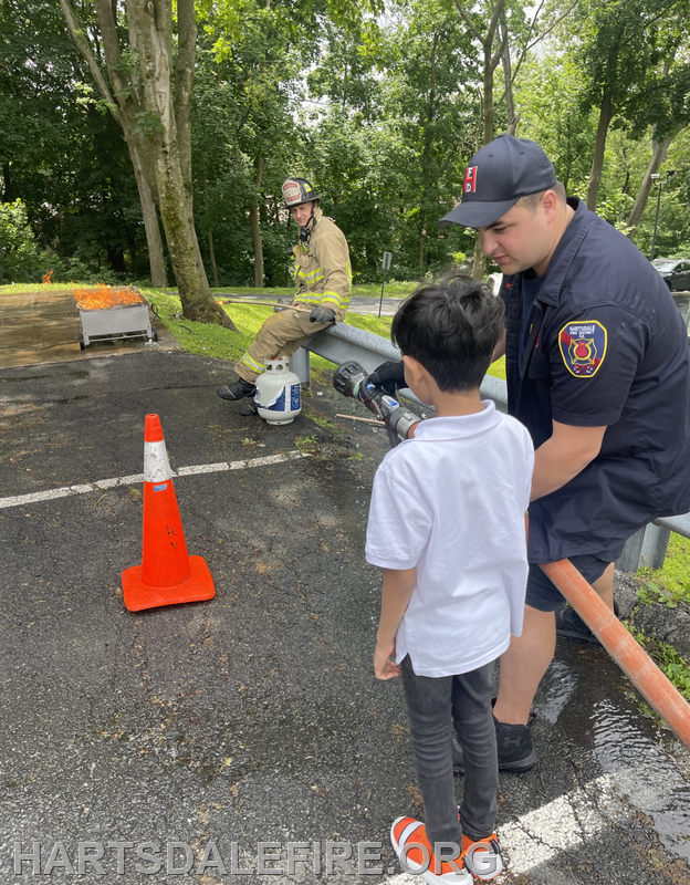 A young child learns to use a fire hose while a firefighter supervises, with another firefighter seated nearby.