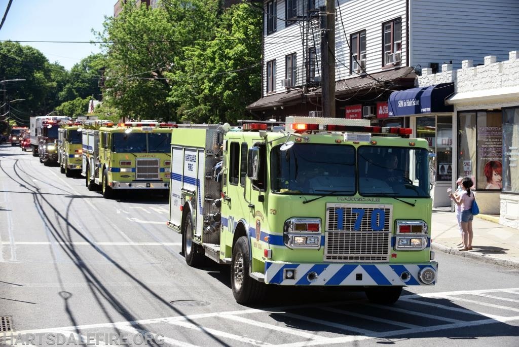 A line of green fire trucks driving down a street with shops and a person taking photos.