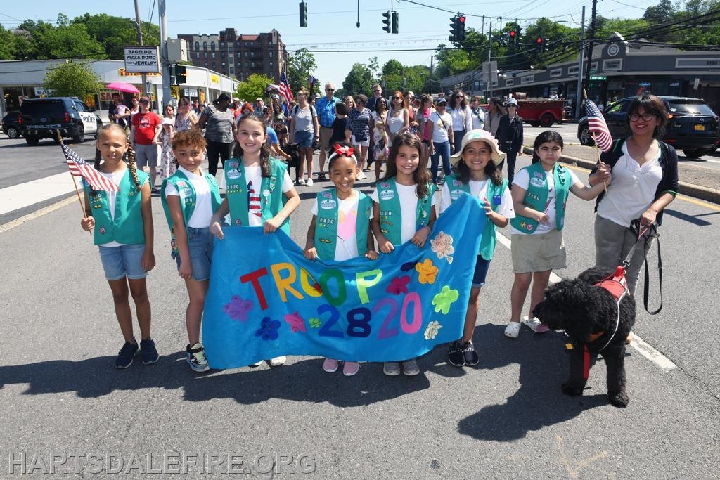 A group of children in green vests hold a "TROOP 2820" banner, standing on a street during a parade.