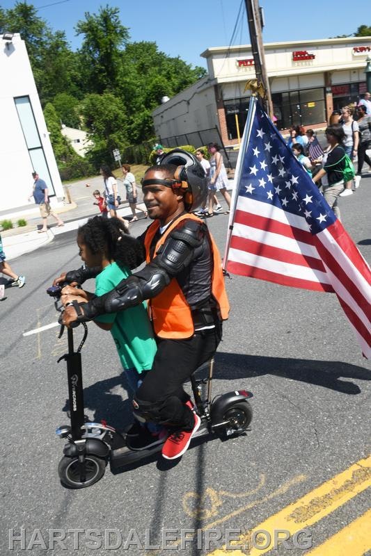 Man and child on a scooter with a U.S. flag in a parade setting.