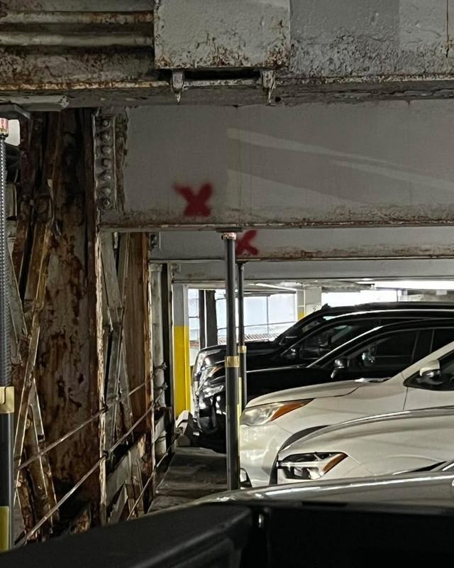 The image shows a parking garage with several cars and some structural elements, including a support pole and rust marks.