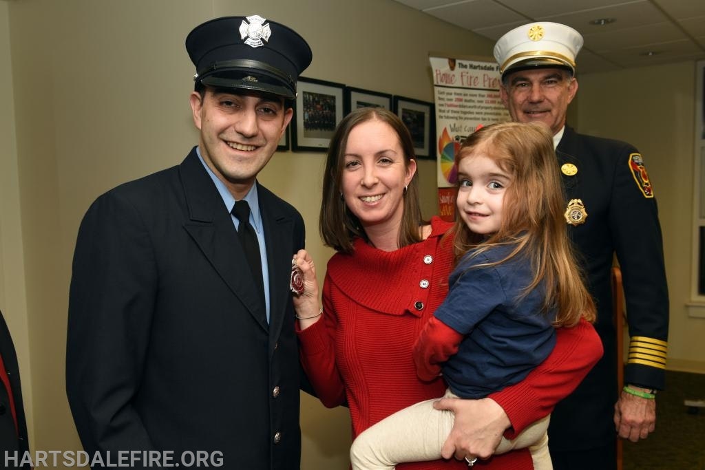 A smiling group with two adults in firefighter uniforms, a woman holding a child, posing indoors with a fire department banner in the background.