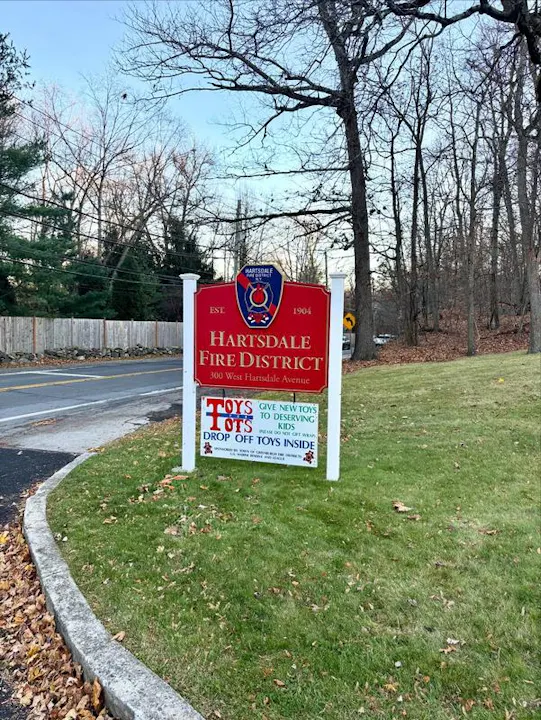 The image shows a sign for the Hartsdale Fire District, promoting a Toys for Tots donation drive at their location.