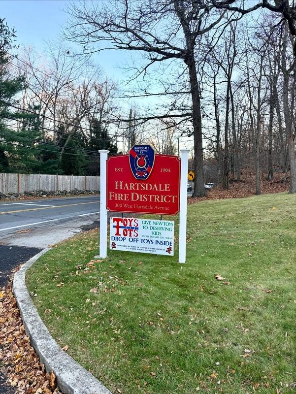 The image shows a sign for the Hartsdale Fire District, promoting a Toys for Tots donation drive at their location.