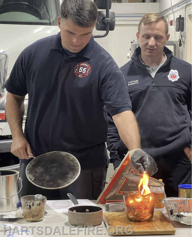 Two firefighters demonstrate a fire safety exercise, featuring flames in a glass container and safety equipment.