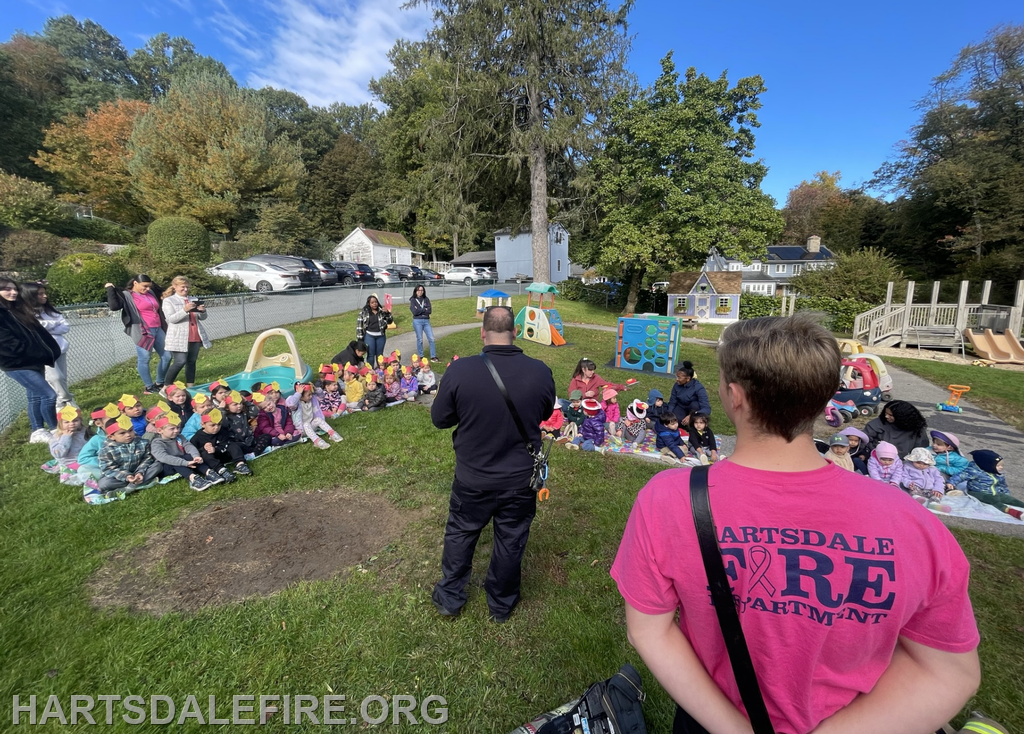 Children in colorful hats sit on the grass, watching a speaker; adults and playground equipment are present in the background.