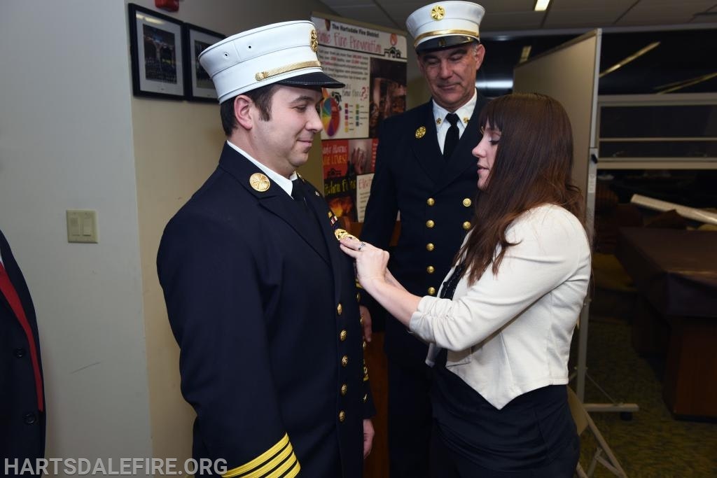 A woman pins a badge on a firefighter in uniform while another firefighter observes.