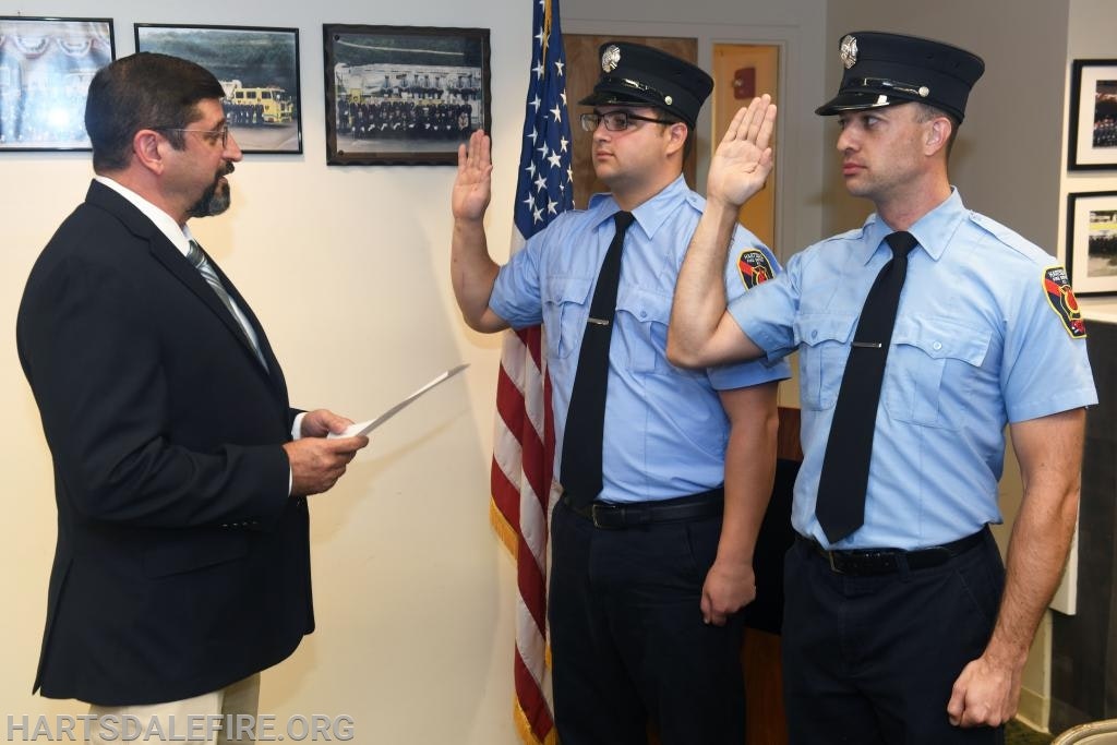 A ceremony with two uniformed firefighters taking an oath, next to an official holding a document, with an American flag in the background.