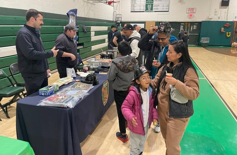 A community event with people enjoying food and activities at a table, kids interacting, and a gym setting in the background.