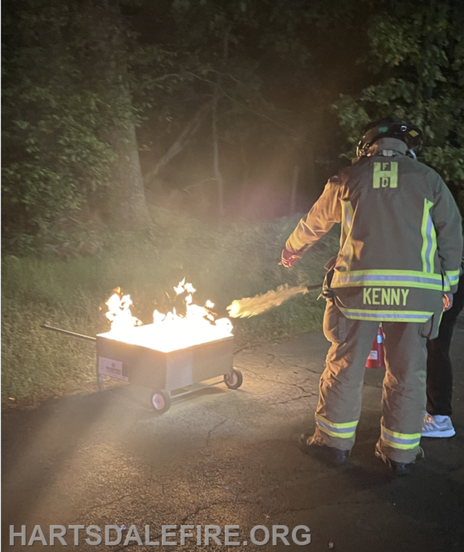 A firefighter in gear uses a tool to manage flames coming from a metal cart in a dark outdoor setting.