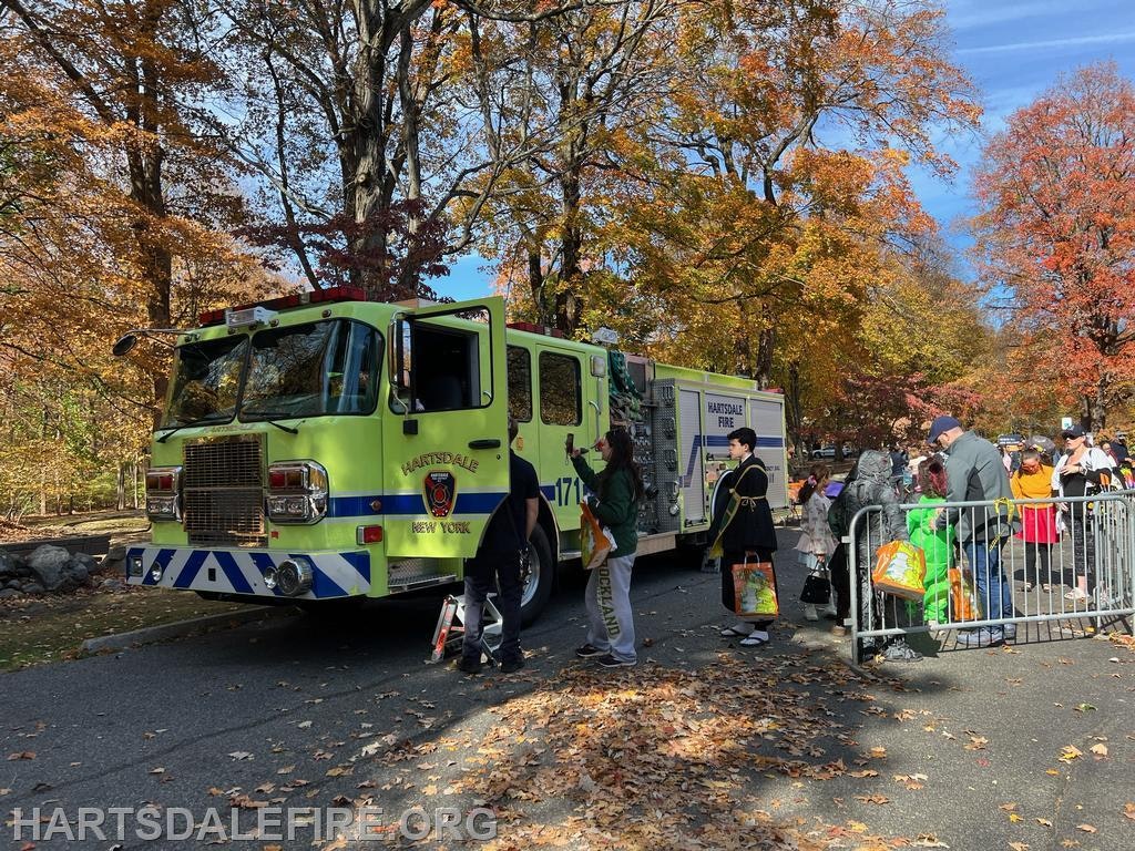 A green fire truck in a park with people, many in costumes, gathering for an event among autumn trees.