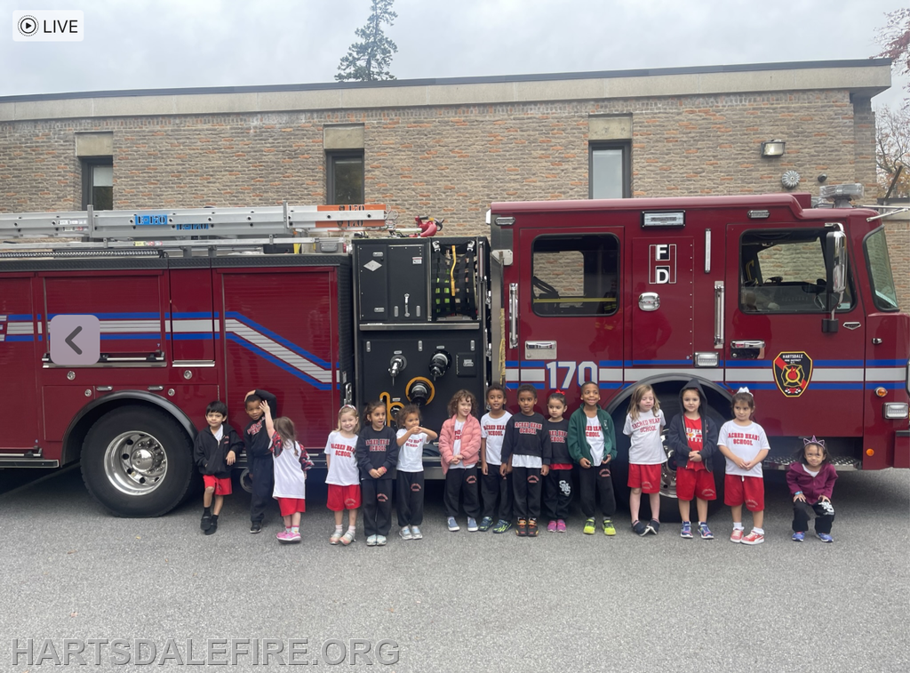 A group of children posing in front of a fire truck, wearing matching shirts, in a school setting.