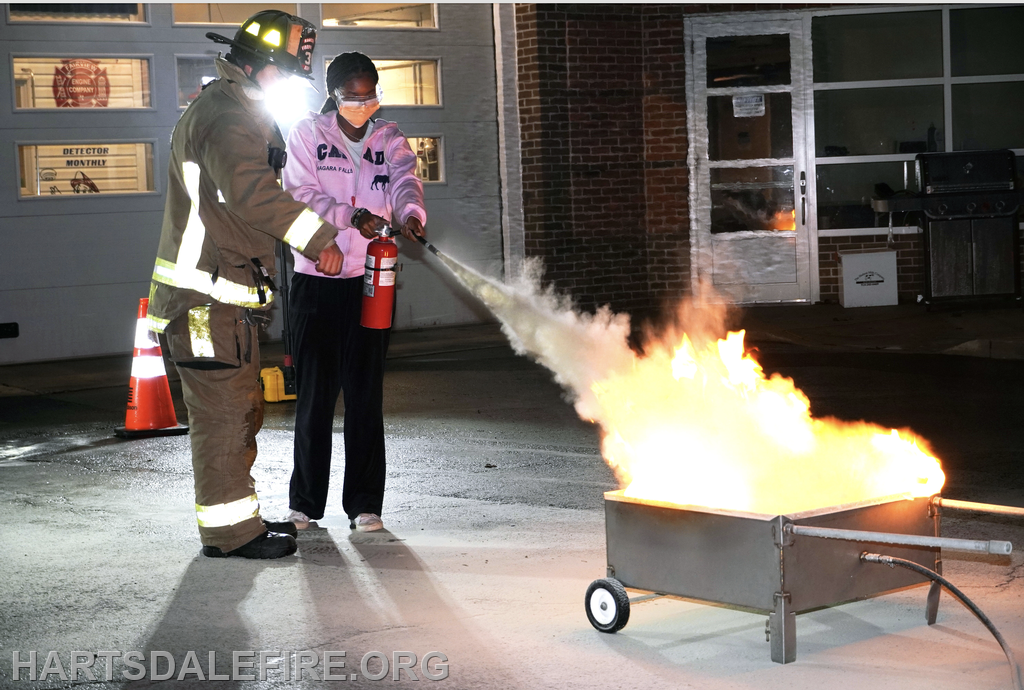 A firefighter demonstrates using a fire extinguisher on a small fire while someone practices under guidance.