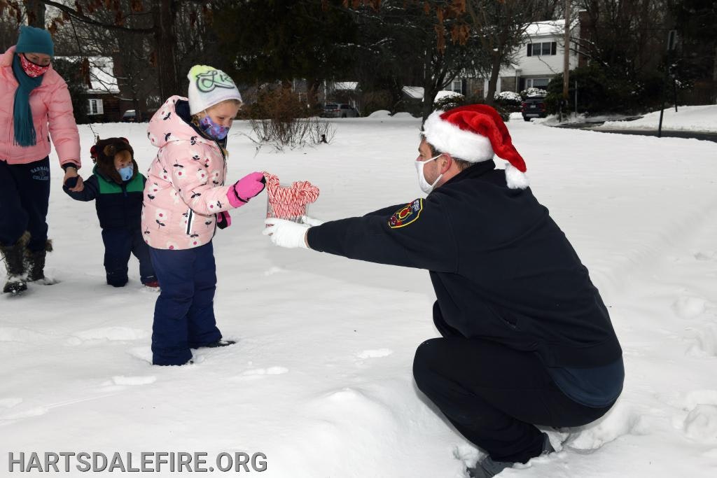 A firefighter in a Santa hat gives a stuffed heart to a child in the snow.