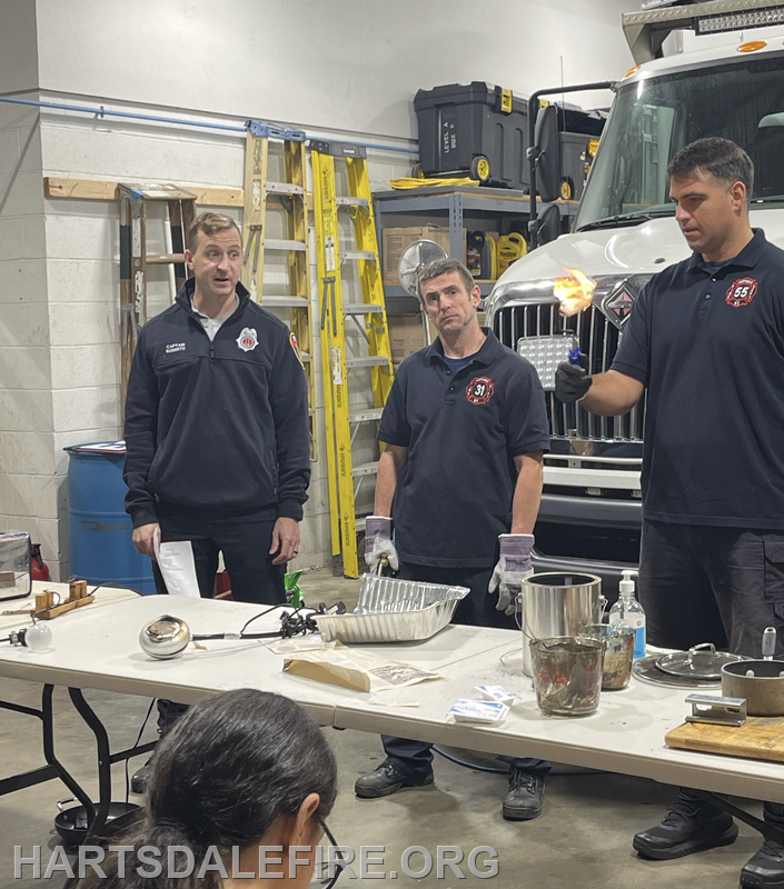 Firefighters demonstrating safety techniques in a training session, with equipment and materials on a table.