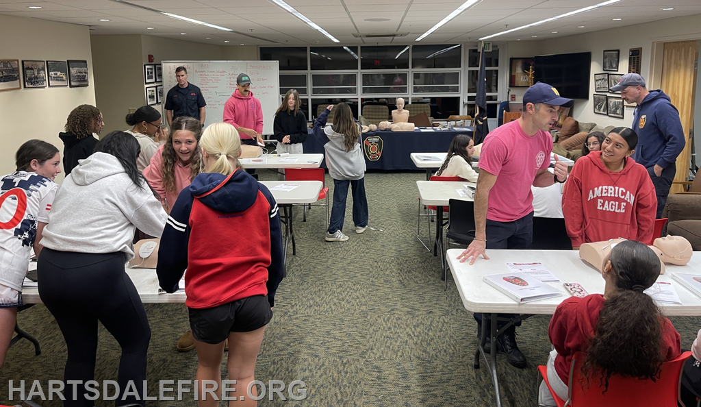 A group of young people participates in a CPR training session, with instructors and practice mannequins in a community space.