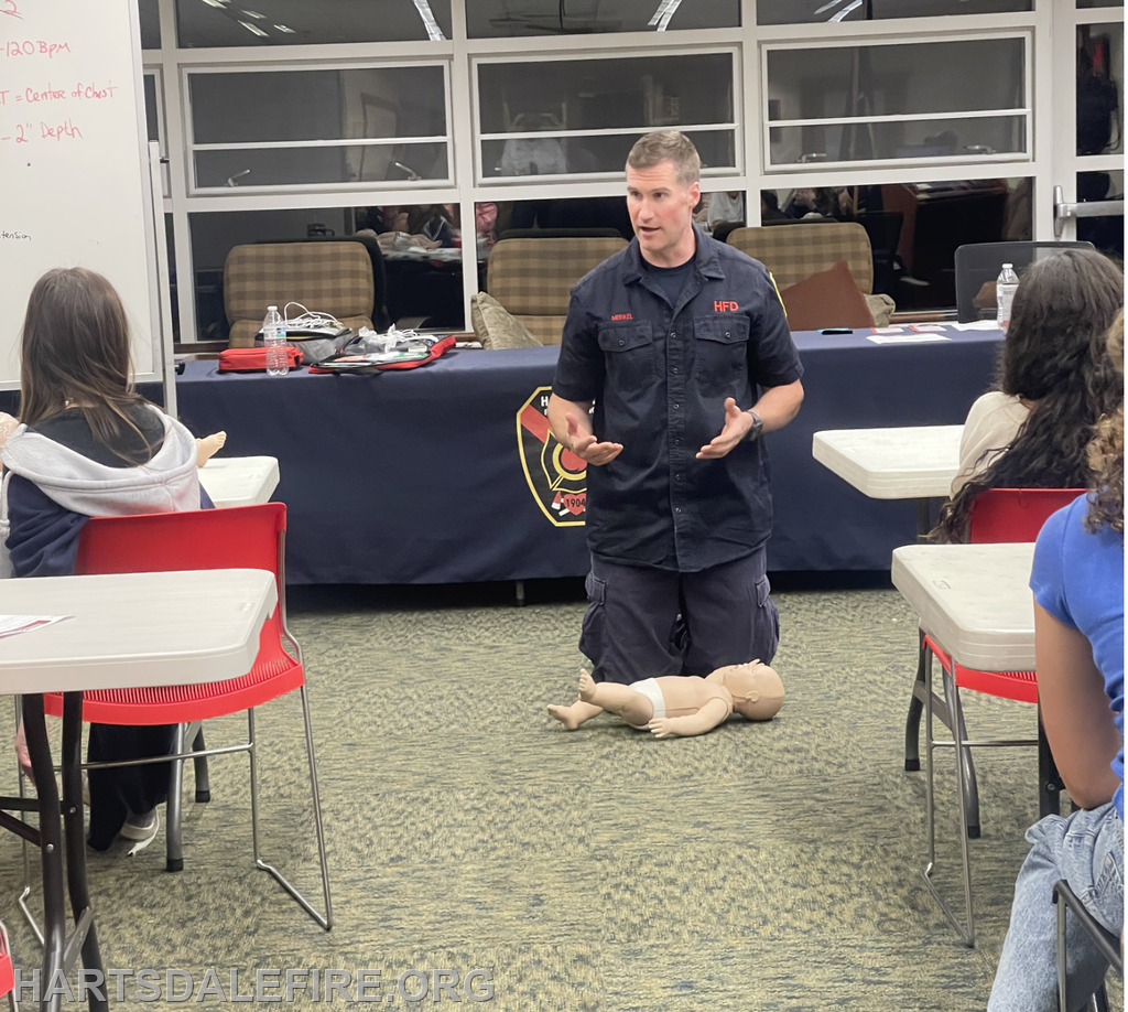 A trainer demonstrates CPR techniques to a group using a mannequin in a classroom setting.