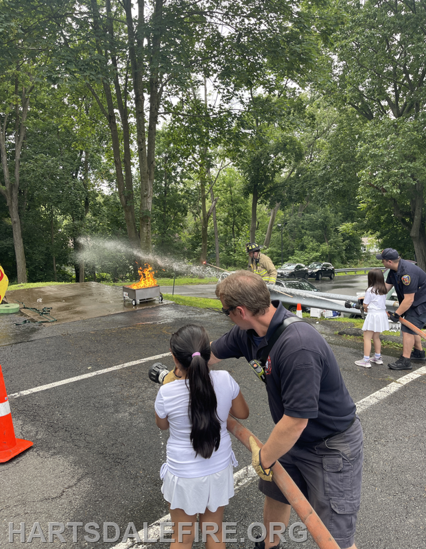 A firefighter demonstrates putting out a small fire while a child observes and handles a fire hose.