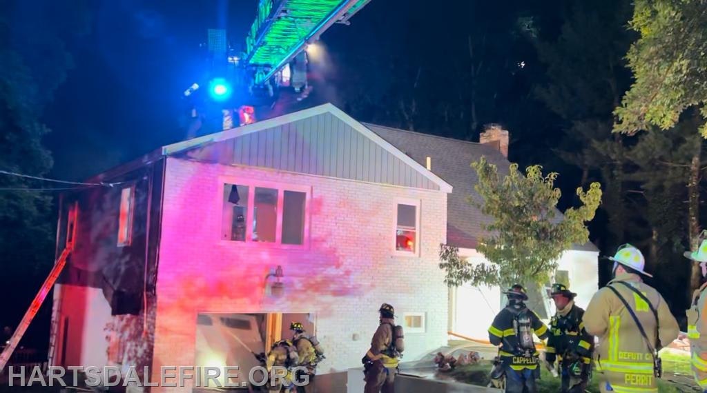 Firefighters responding to a residential fire at night, using a ladder to assist with the situation. Bright lights and smoke are present.