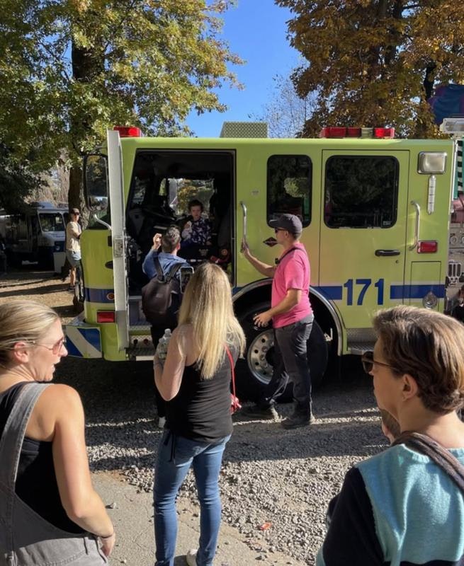 A green fire truck is parked; people are gathered around, interacting, while a child sits inside. It's a sunny day with autumn foliage.