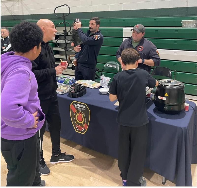 A group chats at a Hartsdale Fire District table, serving food and engaging with children in a gym setting.