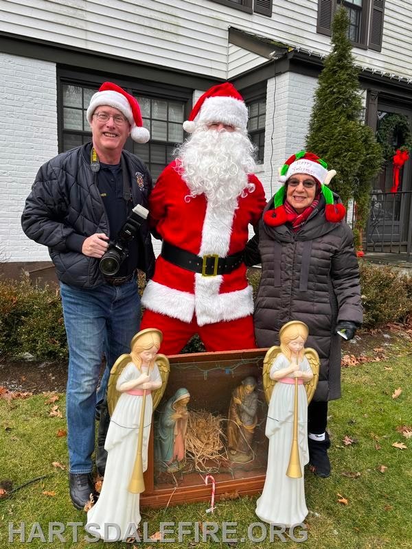 A man with a camera and two people, one in a Santa suit and another in festive attire, pose by a nativity scene decoration.