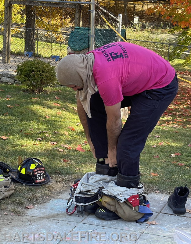 A person in a pink shirt is putting on boots near firefighter gear in an outdoor setting with autumn leaves.