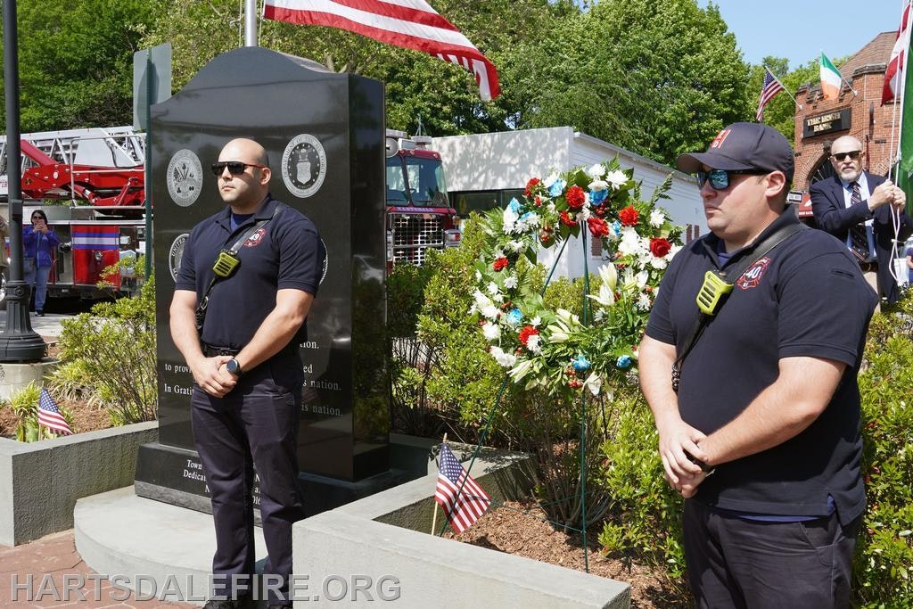 Two uniformed men stand beside a memorial and wreath, with flags and a fire truck in the background.