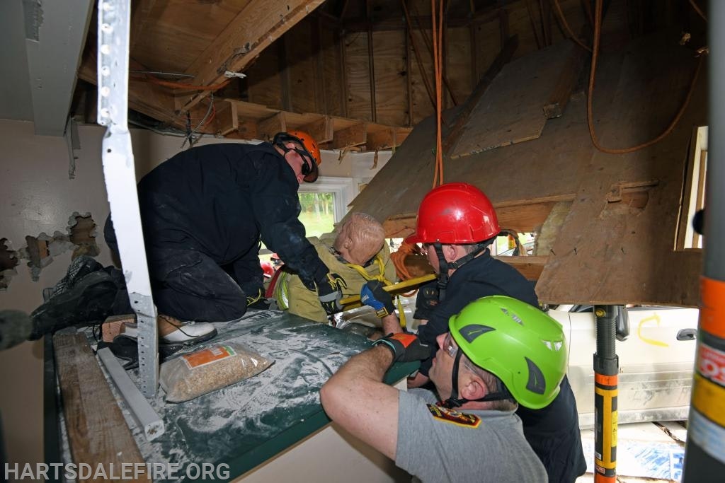 Rescue workers in helmets assist a person in an interior space with damaged walls and ceiling.
