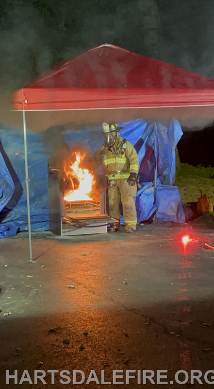 A firefighter in gear stands beside a small fire under a red canopy, with a blue tarp in the background.