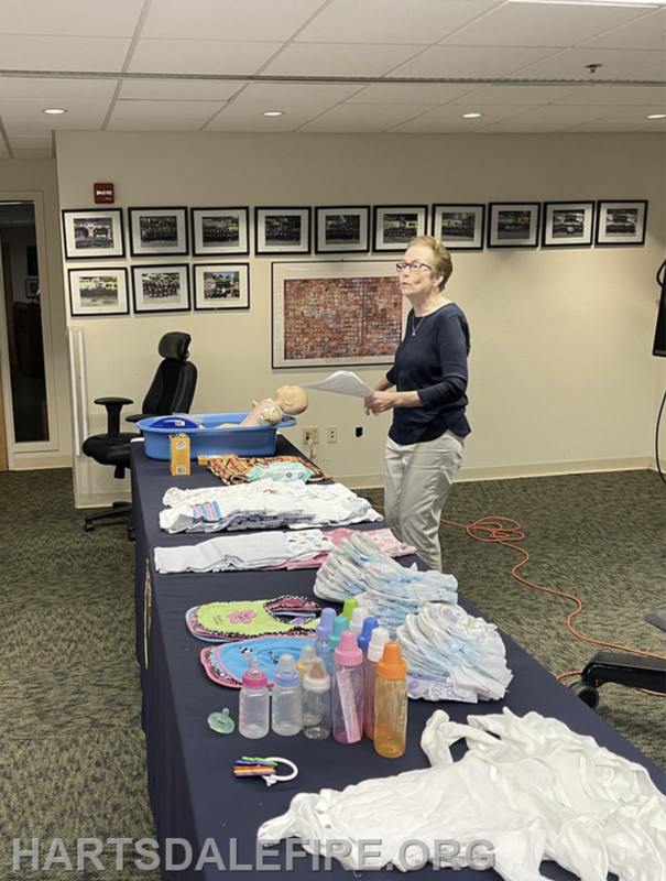 A woman presents in front of a table with baby clothes, bottles, and a doll, likely for a childcare or parenting class.