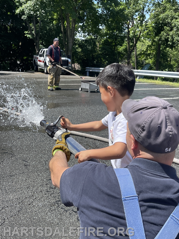 A young boy is learning to use a fire hose with the help of an adult, while another person is nearby. It's a fun fire safety activity!